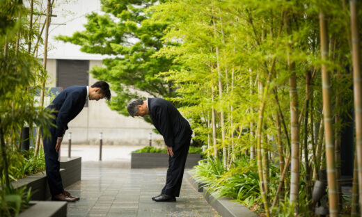 Two business men bow to each other respectfully in a bamboo-lined avenue.