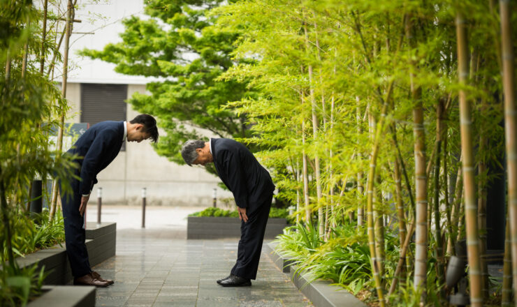 Two business men bow to each other respectfully in a bamboo-lined avenue.